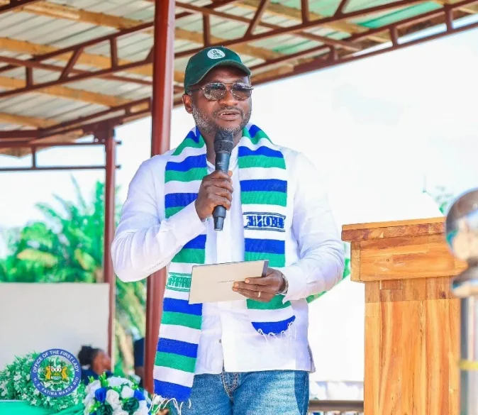 Man in white shirt and green cap speaks into a microphone on a wooden podium, wearing a blue-green striped scarf outdoors.”