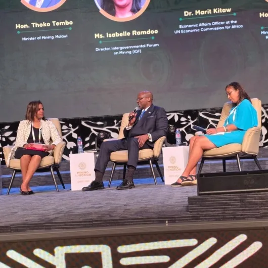 Three panelists seated on stage in beige chairs during a conference talk; the man in the center holds a microphone while two women listen, with bios projected on a large screen behind them.