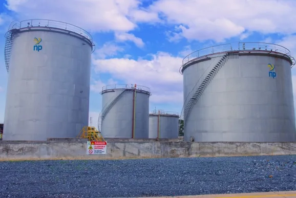 Three large silver storage tanks with external staircases at an industrial facility, blue sky above and a safety sign on the concrete wall in front.